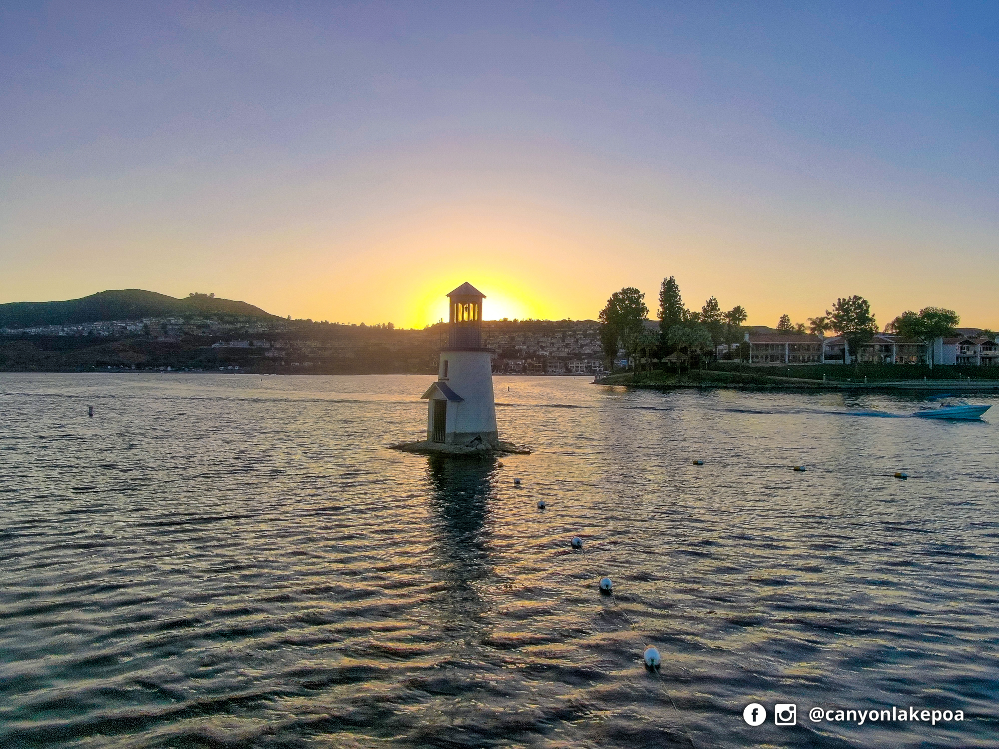 The Canyon Lake lighthouse silhouetted against a warm sunset, with waterfront homes and lake water beyond.
