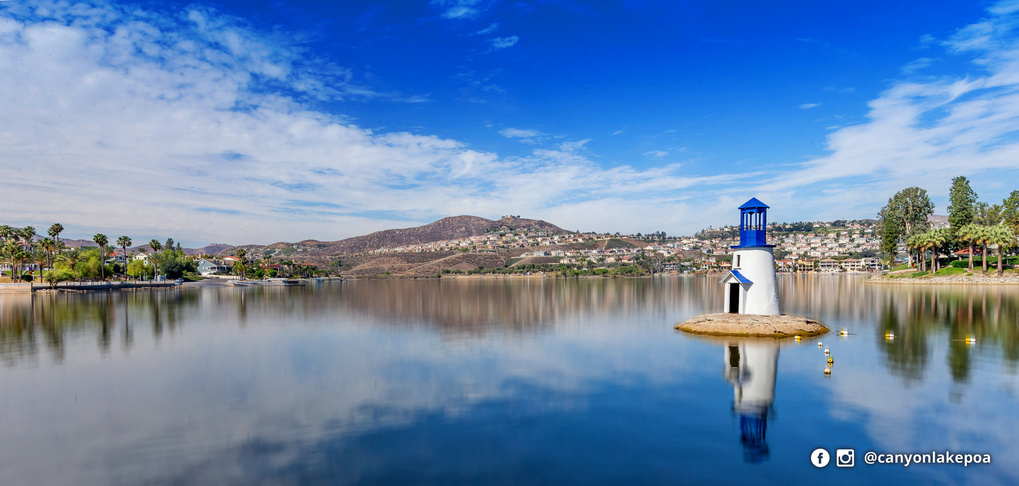 Canyon Lake’s white lighthouse with a blue roof, reflected in calm water, with shoreline homes and hills beyond.